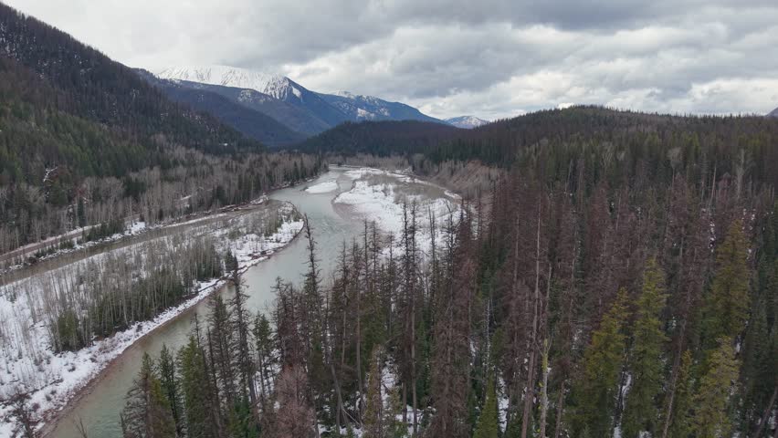 Aerial drone footage of a frozen river winding through a forested mountain valley with snow-covered banks and evergreen trees under a cloudy sky in Glacier National Park, Montana.