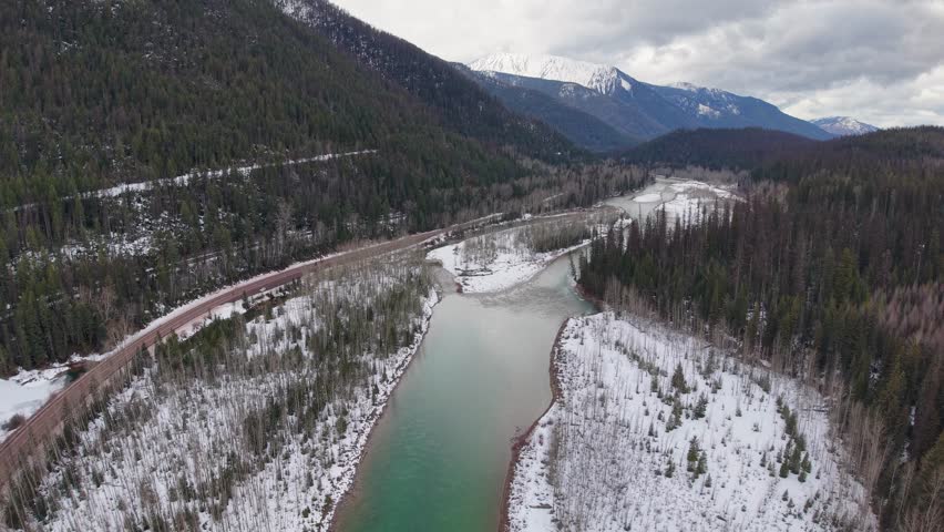 Aerial drone footage of a winding river running alongside a forest road through a snow-dusted valley with evergreen hills and distant mountains in Glacier National Park, Montana.