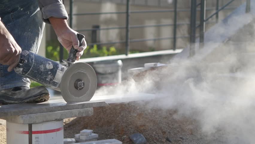 Professional worker cutting large stone tile using electric circular angle grinder with dust clouds