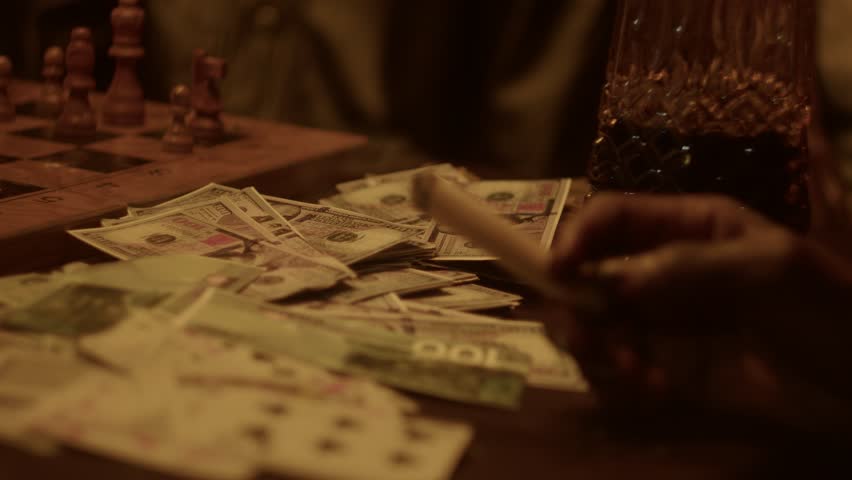 Cinematic close-up of a hand holding a lit cigar over a messy pile of dollars and Ghana Cedi cash, playing cards, and chessboard. Moody, dark lighting, underground gambling, wealth, and power themes.