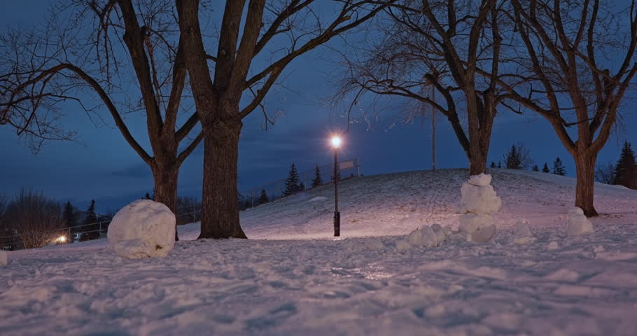 Ground-level view of a quiet winter park evening under warm lamppost light, with snowy ground and bare trees. A small worn snowman sits in the background as the camera glides sideways.