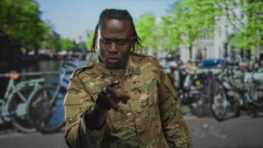 Man soldier in camouflage uniform with hand to neck and dogtag visible on a street by an amsterdam canal; pride duty.