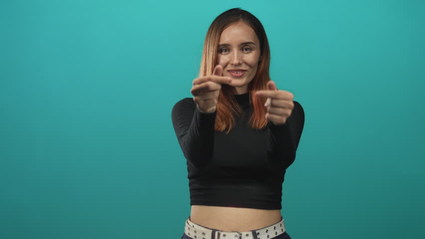 Woman redhead smiling makes finger heart gesture with both hands, showing fingers and bare midriff in a teal studio set wearing black crop top and studded belt; playful affection.
