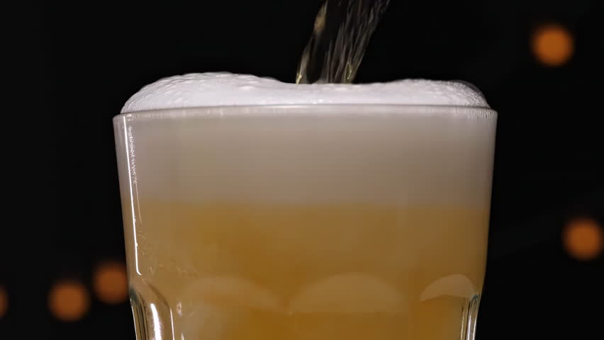 Beer splash - wine, champagne, beer pouring into a glass on dark background with lamps, foam bubbles rising and overflowing, refreshing splash on a black background