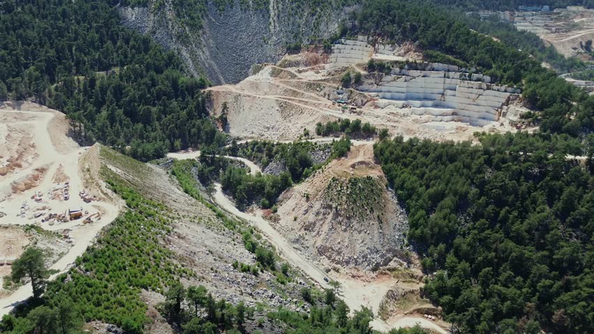 Breathtaking aerial drone shot of an open-pit white marble mine located on a mountain slope next to the turquoise sea. Cinematic view of industrial stone extraction process in a beautiful coastal land