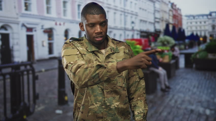 Soldier man in camouflage performing finger to lips for silence and pointing gesture on a street in a city; vigilance duty.