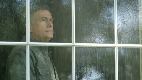 A pensive mature man watches a summer rain shower from his living room window.