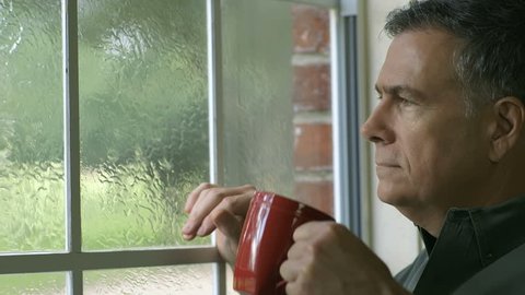 A handsome mature man enjoys a cup of coffee as he watches a summer rain shower through his window.