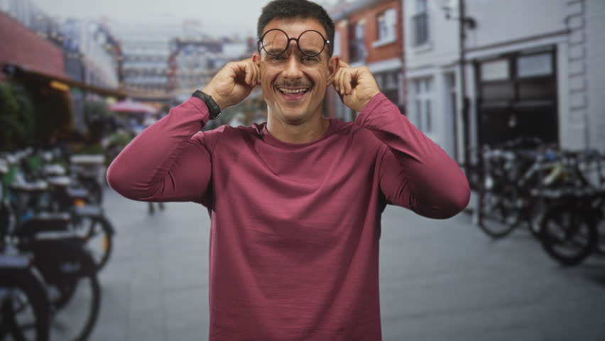Young hispanic man with moustache and round glasses tugs his ears with fingers, smiling in a city street with parked bicycles and storefronts; playful joy.