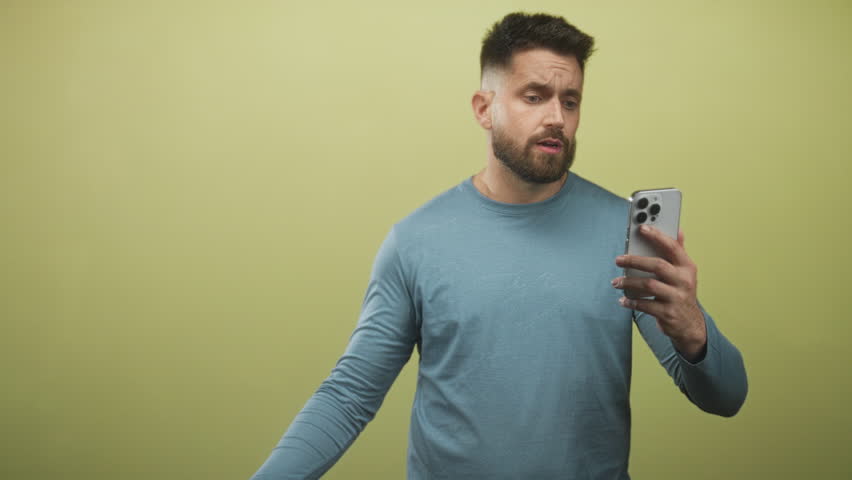 Man holding smartphone in right hand, furrowing brow and looking at screen, wearing blue crewneck shirt in studio; concern.