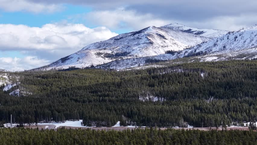 Drone shot of snowy mountain range above dense forest and winding river in Glacier National Park, Montana.