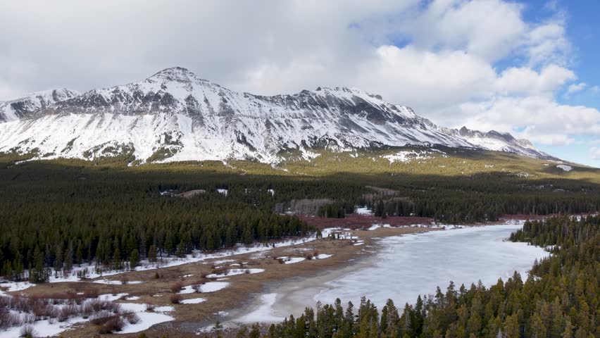 Aerial drone shot over coniferous forest and snowy mountain range in Montana with river partly covered by ice.