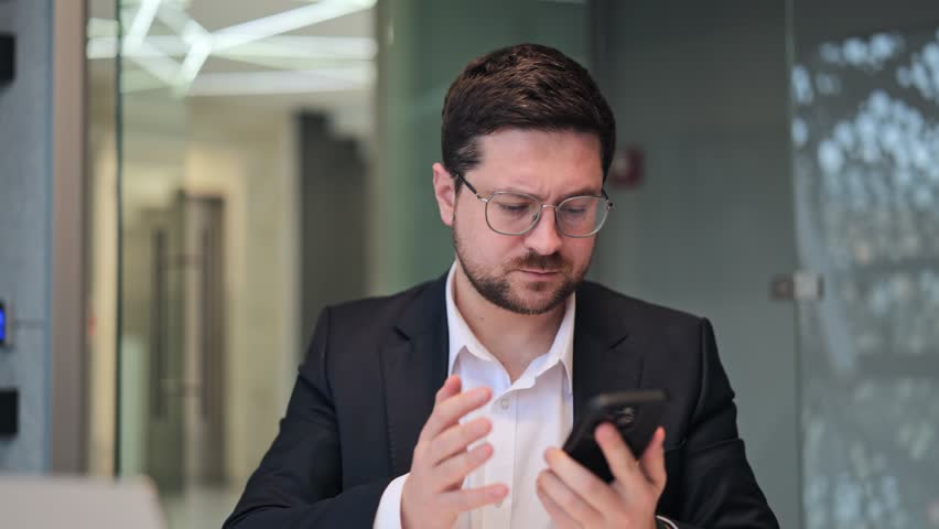 Business professional appears confused while interacting with mobile phone. Moments of frustration and perplexity are evident during problem-solving. Shows modern connectivity challenges in workspace.