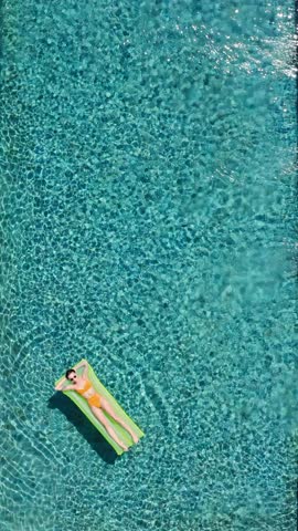 Aerial view of a young woman in orange bikini relaxing and sunbathing floating on a green inflatable mattress in a beautiful turquoise swimming pool water. Vertical Shot