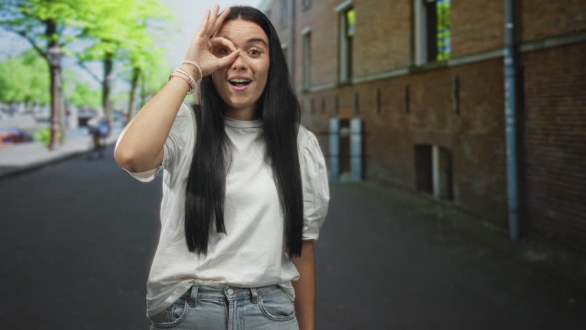 Young hispanic woman standing on a city street by a brick building, making ok sign with hand over eye while smiling in a white t shirt; playful curiosity.