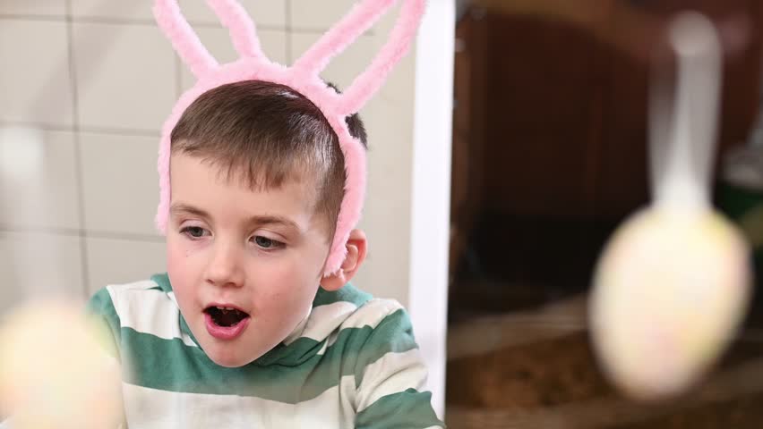 A preschool boy in a bunny costume eats a chocolate egg and a chocolate bunny at home in the kitchen, celebrating Easter