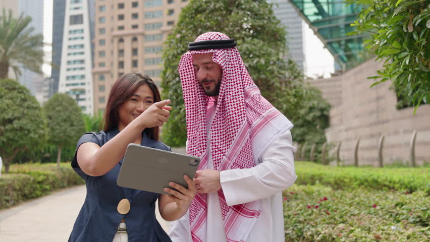 Asian businesswoman and Arab Miiddle eastern businessman talking about cross-border and international business, walking along downtown park.