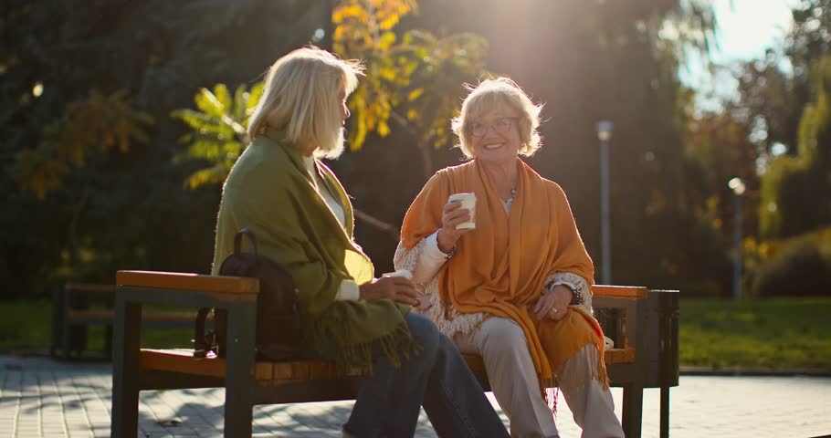 Two elderly female friends sitting on a bench in an autumn park. Cheerful retirees having a friendly conversation while drinking coffee