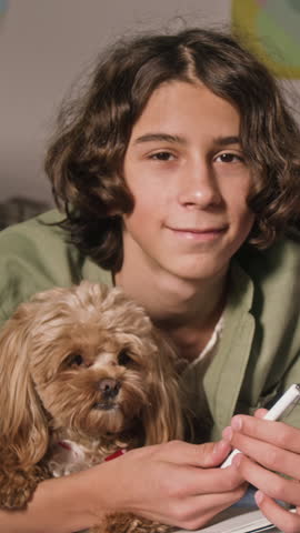 Vertical portrait of cheerful curly-haired teenager smiling for camera holding pen while relaxing on bed with cute sleepy pet