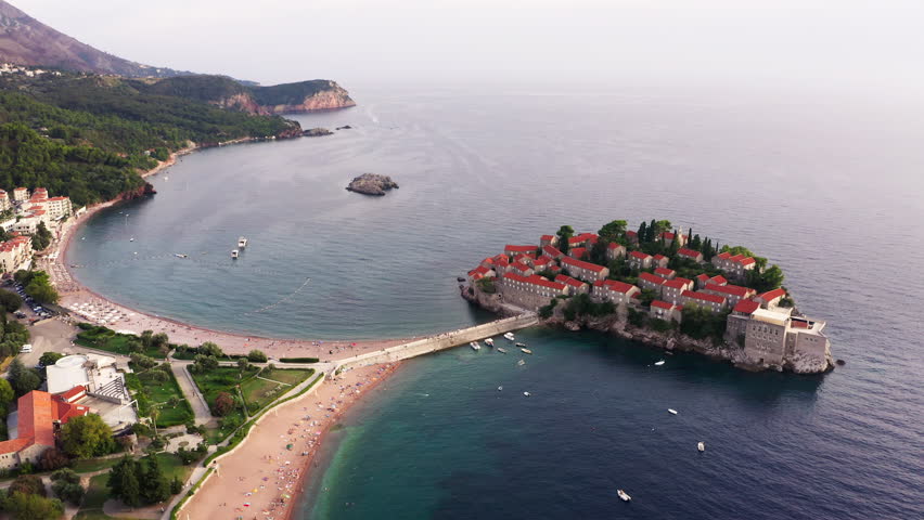 Aerial drone view of Sveti Stefan island in Montenegro. Luxury five star hotel resort on Adriatic Sea peninsula. Mediterranean coastal town landscape with red roofs, blue water and sandy beach. 4K