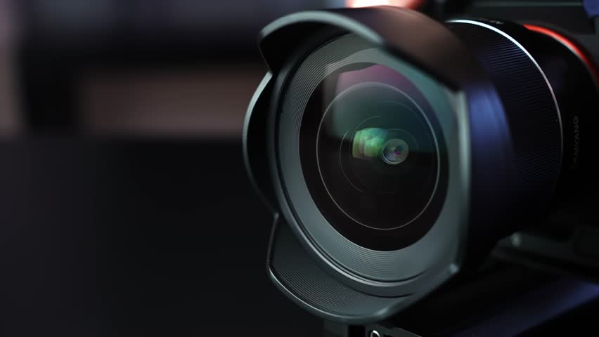 Macro shot of camera lens with hood as aperture slowly opens, revealing shifting reflections in dark studio light with cinematic bokeh—ideal for filmmaking and tech b-roll.
