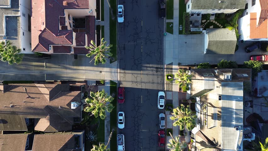 Straight residential corridor near Coronado Ave, Bluff Park shows cracked asphalt, parked cars, palm lined sidewalks in tight coastal grid. Tile roofs, long shadows, warm light, Long Beach, CA. USA