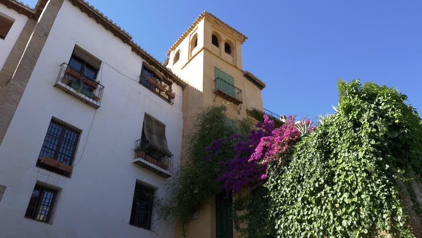 Low angle tracking shot of the historic Casa del Almirante. Features vibrant purple bougainvillea flowers, traditional Spanish architecture, and a clear blue sky in the heart of Granada.