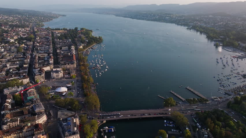 Smooth drone shot orbiting the Quaibrücke in Zurich with sweeping views over Lake Zurich and the city waterfront. Golden light, calm water reflections, and cinematic Swiss urban atmosphere.