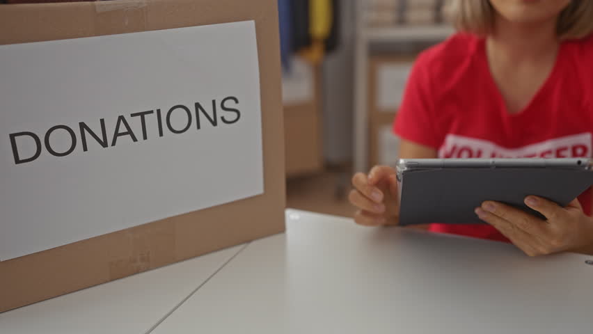 Blonde woman taps tablet screen near a large cardboard donation box at a donation center in building with shelves of clothes; community support.