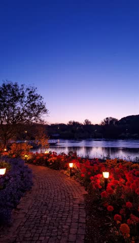 Enchanting night garden path with glowing lanterns and red flowers by a peaceful lake at twilight
