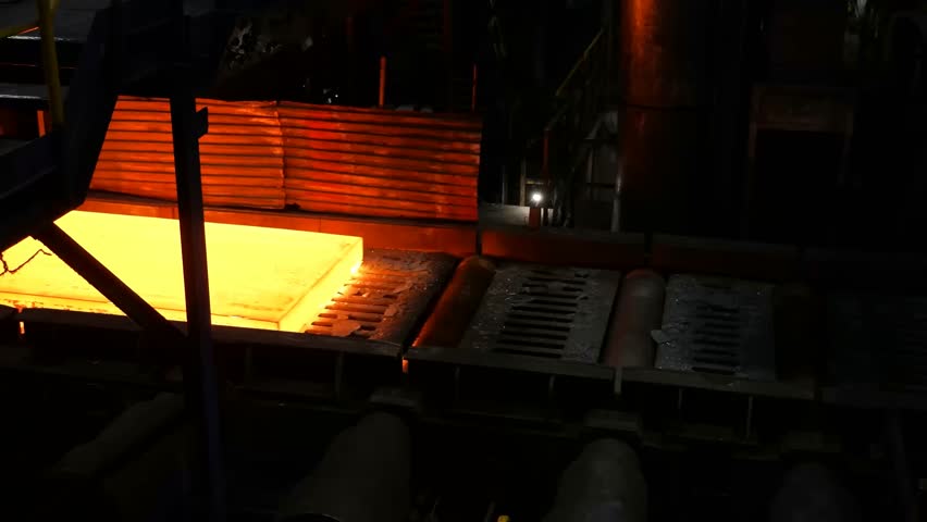 The industrial steel slabs move along heavy rollers in a dark factory while cooling water spray creates massive steam clouds around the glowing metal