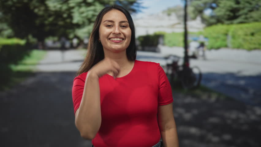 Woman touching cheek with hand wearing red shirt on tree lined street near parked bicycle; joy confidence.