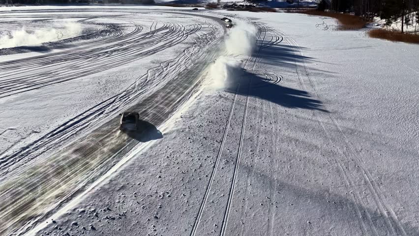 Drone aerial of a car drifting on a frozen lake, throwing a huge snow cloud and leaving tire tracks across the winter ice surface.