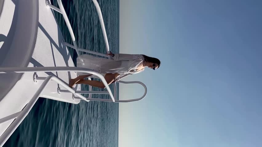 Woman in white dress elegantly moves along boat railing above tranquil ocean water, with sunlight enhancing the peaceful maritime scene