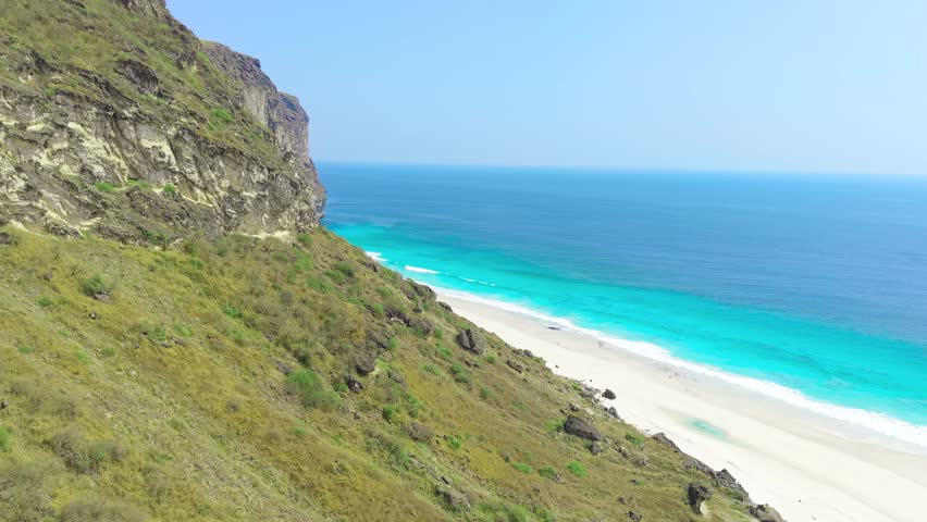 Aerial view of high rocky cliffs bordering a white sand beach and turquoise ocean water on Socotra Island, Yemen.