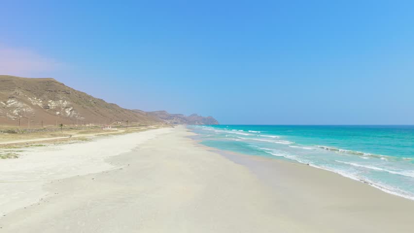 Aerial view of a wide white sand beach with turquoise ocean water and rocky mountains in the background on the island of Socotra, Yemen.