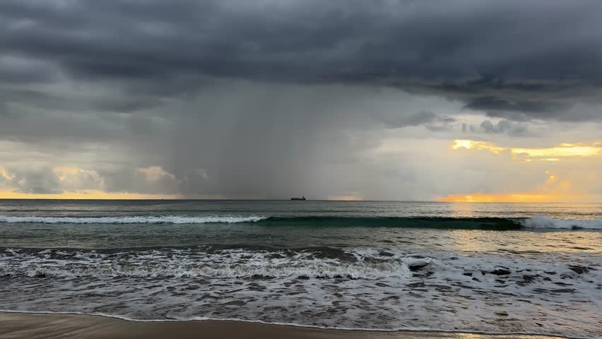 A dramatic sunset view on a tropical beach with dark storm clouds and a visible rain shaft or water column falling from the clouds into the sea.