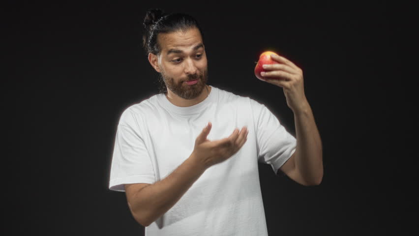 Young arab hispanic man holding red apple with outstretched hand in studio under black backdrop and studio lighting; healthy choice playful.