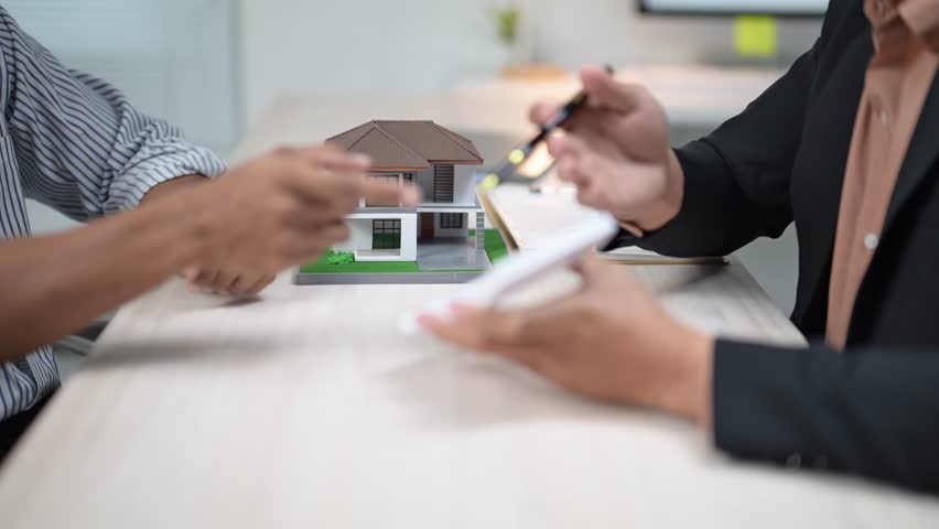 Two people are sitting at a table with a house model in front of them. One of them is holding a pen and the other is holding a remote. They are discussing something related to the house model