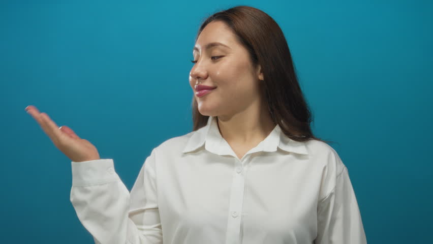 Woman holds out palm presenting gesture with subtle smile wearing white blouse against bright blue studio wall; friendliness.