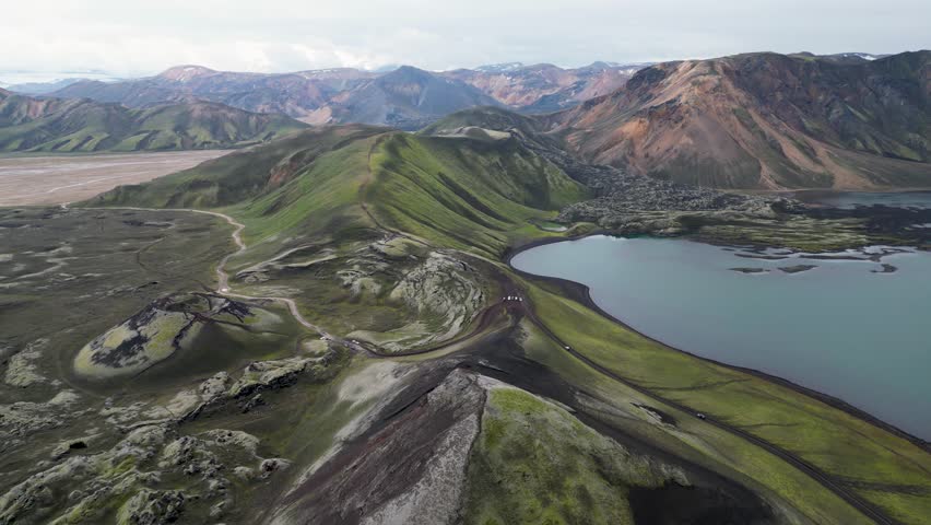 Aerial panoramic view of the colorful rhyolite mountains, volcanic landscape and lake in Landmannalaugar, Iceland. Dramatic highland scenery with green hills, lava fields and winding road in the Icelandic Highlands, popular destination for hiking and adventure travel.