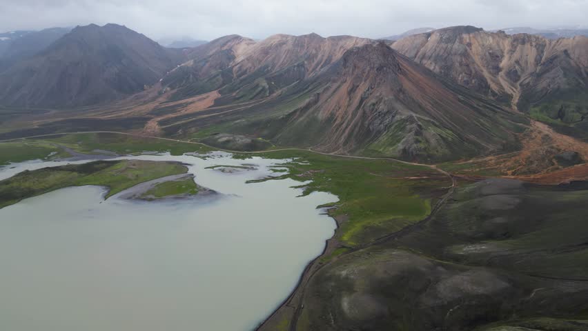 Aerial panoramic view of the colorful rhyolite mountains, volcanic landscape and lake in Landmannalaugar, Iceland. Dramatic highland scenery with green hills, lava fields and winding road in the Icelandic Highlands, popular destination for hiking and adventure travel.