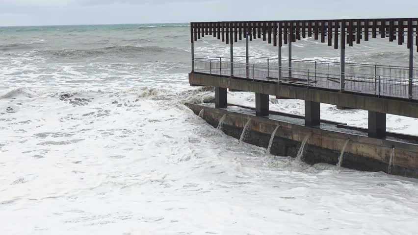 Raging ocean crashes structure violently. Stormdriven waves batter harbor defenses intensely. Powerful storm surge causes flooding and aggressive wave activity visualized