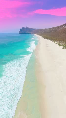 Aerial view of waves breaking on a white sand beach along turquoise ocean waters under a pink sunset sky at Socotra, Yemen.
