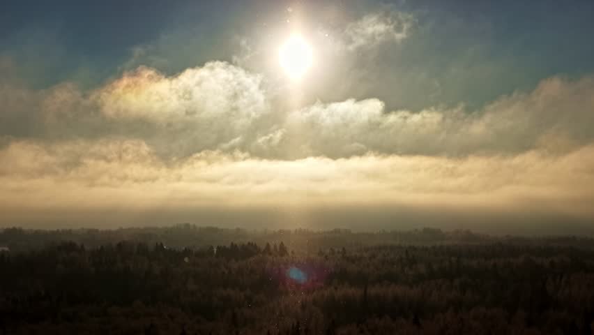 A bright sun above white clouds over a winter landscape during a light snowfall.