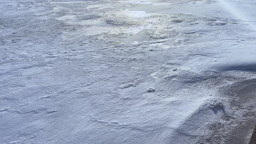 Panoramic view of a wide frozen river covered with textured ice and snow under bright winter sunlight with natural lens flare.