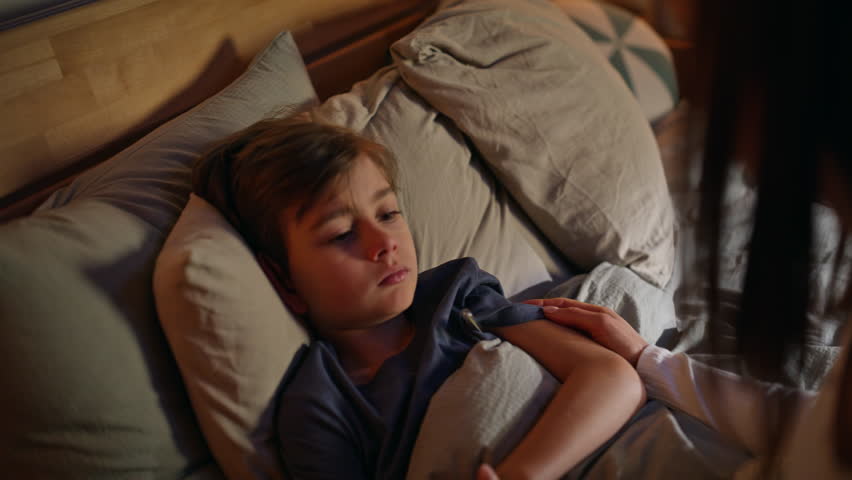 Mother hands checking thermometer caring health little boy lying comfortable home bed late evening closeup. Caring young mom touching son forehead measuring temperature at bedtime. Domestic treatment.