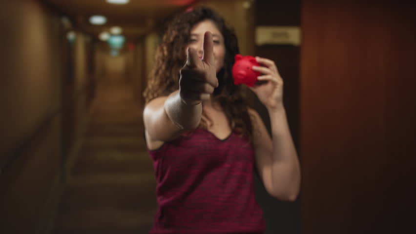 Young hispanic woman points finger forward while holding a red piggybank in a hotel building; confidence savings.