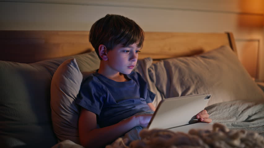 Boy browsing tablet night bedroom closeup. Little kid focused pad computer touching screen watching online content lying cozy bed alone. Addicted child using digital gadget at bedtime. Childhood habit