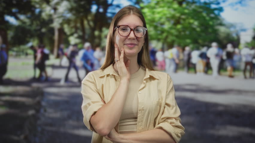 Young blonde woman smiles while touching cheek in sunlit green park outdoors with blurred crowd walking behind; serenity.